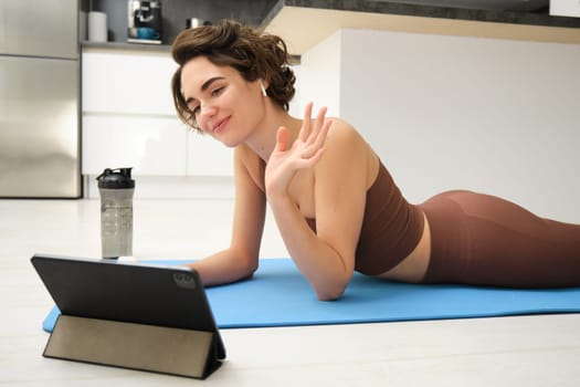 Sport and lifestyle. Young fitness woman, gym instructor waves hand at tablet, joins online video class, yoga and mindfulness workout, training from home indoors.