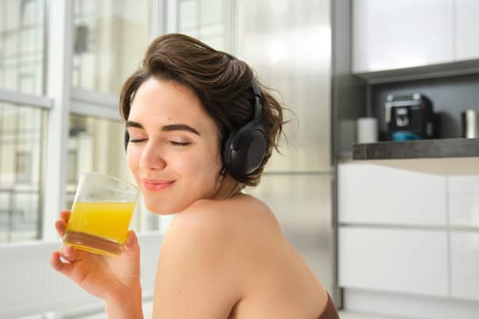 Smiling sportswoman, fitness girl does workout at home, wears headphones, listens music and drinks orange juice after training session.