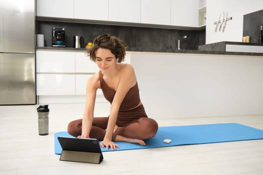 Portrait of smiling young woman, fitness instructor on rubber yoga mat, joins online training class from tablet, listens video in wireless headphones, workout at home on floor.