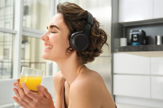 Sport and healthy lifestyle. Portrait of happy sporty girl, fitness woman in headphones, rewinds at home, drinks orange juice with happy smile after workout.