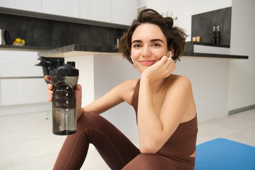 Portrait of fitness woman, young sportswoman at home, drinks water from bottle after workout, training exercises, takes a break after pilates, yoga training, stays hydrated.