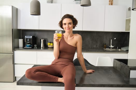 Beautiful smiling fitness girl, wearing activewear, sitting on kitchen counter, drinking glass of orange juice and looking happy. Sport and healthy lifestyle concept