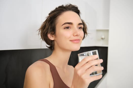 Close up portrait of smiling fitness woman, holding glass of water and lookins away pleased. Girl drinks after workout. Sport and healthy lifestyle concept