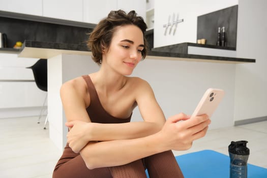 Portrait of sporty girl with mobile phone, sitting on yoga mat, using smartphone during workout at home, watching fitness turorial on telephone. Sport and wellbeing concept