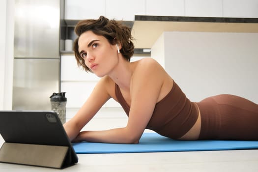 Portrait of fitness girl with thoughtful face, lying on rubber yoga mat at home, listening and watching sport workout video tutorial on tablet, thinking.