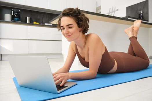 Portrait of beautiful sportswoman, fitness girl watching videos on laptop during workout, follow online pilates videos while exercise at home, wearing activewear in kitchen.