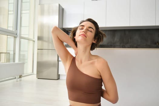 Young smiling brunette woman, yoga girl stretching her arms behind back with pleased, relaxed face expression, workout in bright room at home.