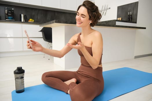 Portrait of happy smiling, relaxed fitness instructor, girl video chats, looks at smartphone, records workout blog, yoga online, sits on sports mat in kitchen, does pilates from home.