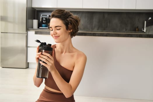 Fitness instructor, smiling young woman workout at home, sits on yoga mat and drinks water, does exercises, training session in her kitchen. Sport indoors concept