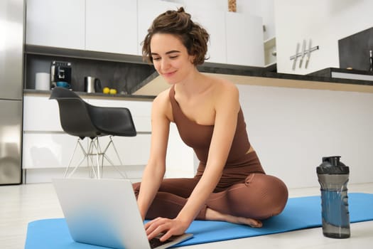 Portrait of young female athlete, fitness instructor starting online gym class from her home, sitting on rubber yoga mat, typing on laptop. Sport and lifestyle concept
