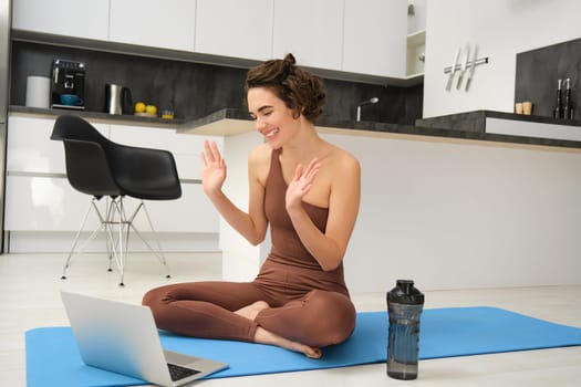 Young smiling woman sits on sports rubber mat in activewear, says hello at video chat online yoga class, using laptop during home workout training session. Copy space