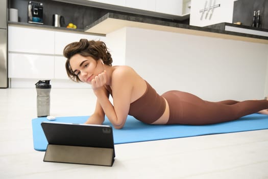 Young fitness woman, girl in tracksuit lying on fitness mat, looking at yoga, pilates video tutorial on tablet, does workout from home, uses gym app, training at home.