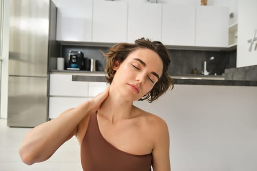 Close up portrait of sportswoman, fitness woman massaging her neck, warm-up her body after workout, doing yoga on rubber mat at home.