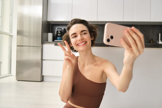 Smiling fitness girl, takes selfie during workout at home, posing for photo and holding smartphone, workout from home, recording vlog of her yoga training session.