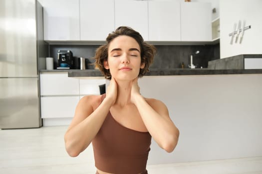 Close up portrait of sportswoman, fitness woman massaging her neck, warm-up her body after workout, doing yoga on rubber mat at home.