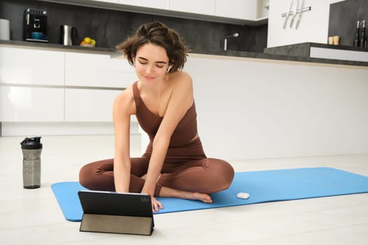 Portrait of smiling young woman, fitness instructor on rubber yoga mat, joins online training class from tablet, listens video in wireless headphones, workout at home on floor.