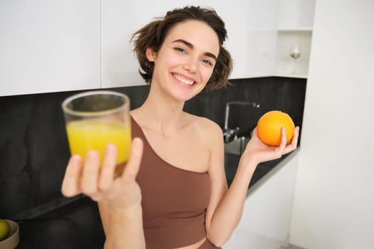 Healthy body and wellbeing. Smiling fitness girl giving you glass of fresh juice, holding an orange and looking happy. VItamin drink after workout at home.