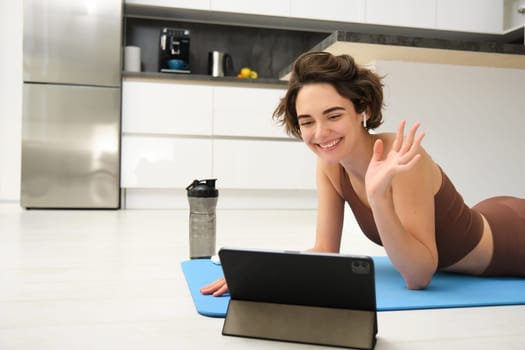 Smiling brunette woman in sportswear, waves hand, says hi at tablet, records video sports blog, fitness training session online, instructor shows workout elements, uses rubber mat at home in kitchen.