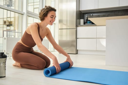 Healthy lifestyle and sport. Fitness woman roll out her yoga matress for workout at home, prepare water bottle. Girl does sports indoors, wears activewear, prepares for training session.
