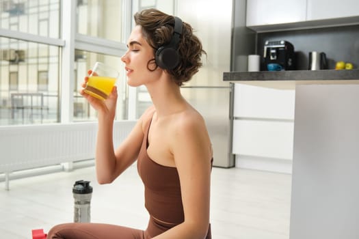 Sport and healthy lifestyle. Portrait of happy sporty girl, fitness woman in headphones, rewinds at home, drinks orange juice with happy smile after workout.