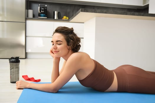 Sport and healthy lifestyle. Portrait of brunette sportswoman doing fitness in activewear, lying on rubber mat on floor, doing workout training in kitchen, drinking water from gym bottle.