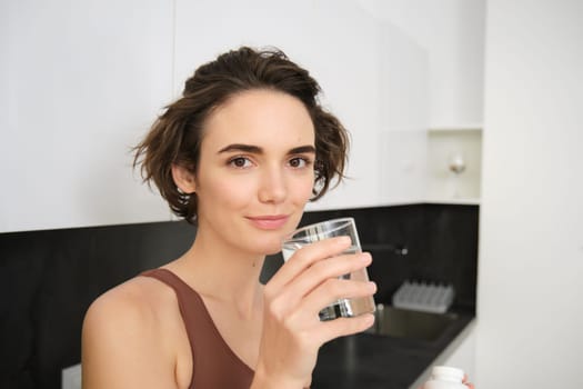 Healthy lifestyle and female wellbeing. Young beautiful woman standing in kitchen and drinking glass of water after workout training. Fitness girl stays hydrated after gym class.