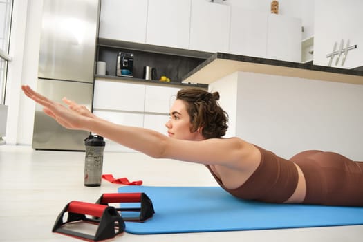 Portrait of sportswoman at home, doing workout training, stretching arms forward, leading active lifestyle from indoors, using resistance band and plunk push up bars for exercising.