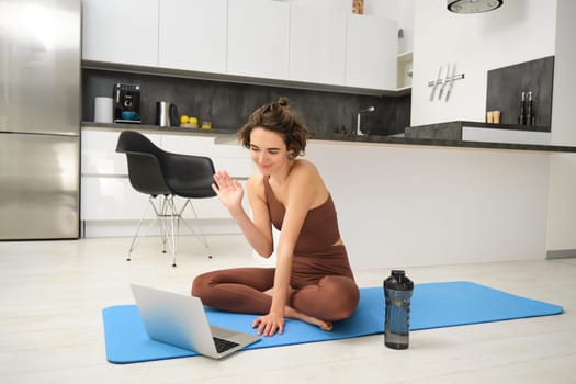 Sport and women. Young fitness instructor, yoga girl sits on rubber mat in her kitchen, waves hand and says hello at laptop, has online video gym class from home.