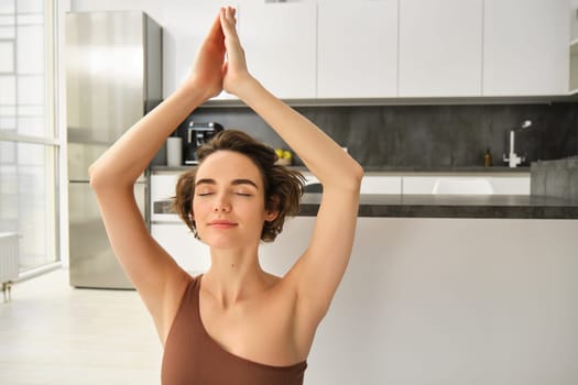 Young calm fit healthy woman wearing sportswear sitting at home in living room doing yoga exercise, meditating and breathing in the morning. Mental health and zen meditation concept.