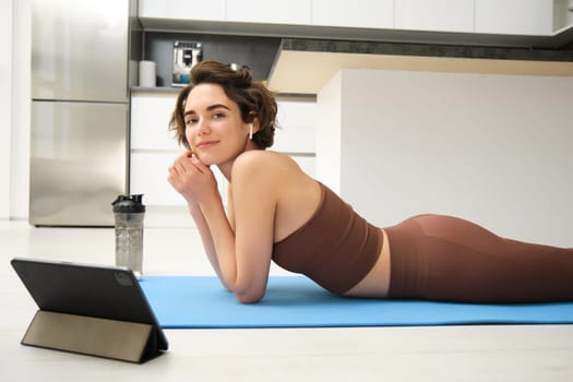 Portrait of beautiful woman in fitness sportswear, lies on floor, rubber yoga mat, uses tablet and wireless headphones, watches online sport workout tutorial, gym classes online from home.