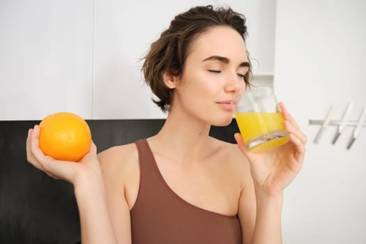 Image of sportswoman, fitness girl holding glass of juice and an orange, smiling, drinking vitamin beverage after workout, standing in her kitchen at home. Healthy lifestyle and sport concept.