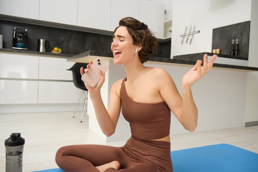 Carefree fitness girl, workout instructor sits at home with water bottle, yoga mat, wears tracksuit, sings into her smartphone, enjoys music in wireless headphones, listens song during pilates yoga.