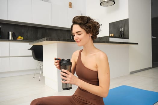 Portrait of fitness woman, young sportswoman at home, drinks water from bottle after workout, training exercises, takes a break after pilates, yoga training, stays hydrated.