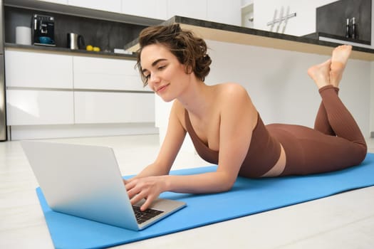 Portrait of woman athlete, girl workout at home, watches fitness videos on laptop, lying on rubber mat and doing indoor yoga session. Sport and wellbeing concept