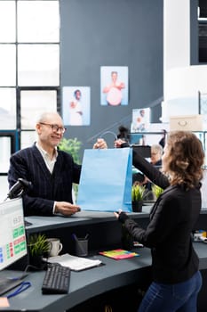 Senior shopper buying fashionable clothes in mall store, making electronic payment at counter desk. Cheerful client talking to store worker discussing casual wear promotions, commercial activity.