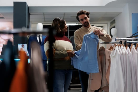 Clothing store customer getting fashion recommendation from consultant while browsing trendy apparel rack. Woman and boutique worker holding shirt on hanger and selecting size