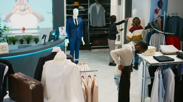 Multiethnic group of people looking at trendy clothes in retail store market, shopping for formal or casual wear. Diverse customers examining fashion collections on hangers, commercial activity.