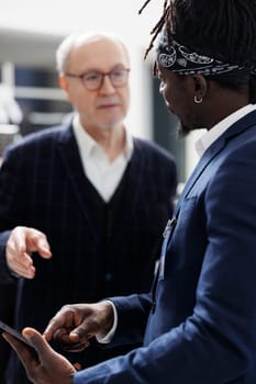 African american worker checking stylish shirt inventory on tablet computer for senior client in shopping centre. Elderly man buying formal clothes and fashionable merchandise in modern boutique