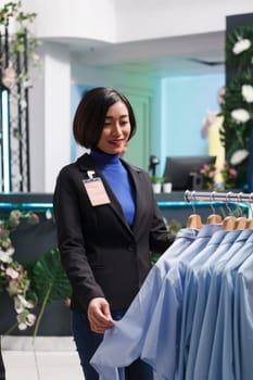 Shopping center clothing store seller exploring formal apparel rack and managing inventory. Fashion boutique asian woman employee standing near apparel rack and checking merchandise