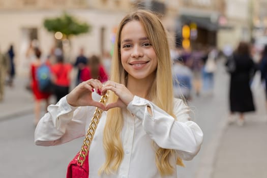 I love you. Young woman makes symbol of love, showing heart sign to camera, express romantic feelings, express sincere positive feelings. Charity, gratitude, donation. Outdoors in urban city street