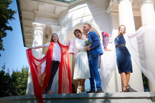 Kirov, Russia - August 10, 2021: Groom and bride walking in park on sunny summer day and bridesmaids with beautiful fabrics accompanying them. Friends and guests at wedding with newlyweds outdoors