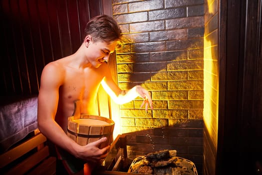 Kirov, Russia - November 10, 2021: Teenage boy in underwear in bathhouse, sauna at spa center with bucket of water and heated stones for steam and warmth. Concept of maintaining health and pleasure