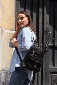 Beautiful young girl wearing blue jeans, shirt and white t-shirt posing with black leather backpack. Street photo