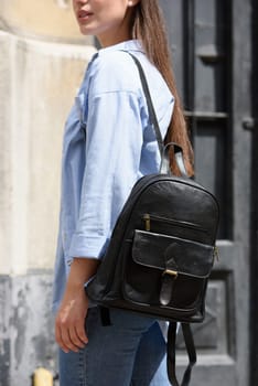 Beautiful young girl wearing blue jeans, shirt and white t-shirt posing with black leather backpack. Street photo