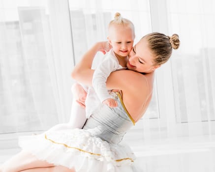 Young mother in swan ballet dress hugging little smiling daughter sitting on the floor in light room. Positive emotions of mother and daughter in ballet studio