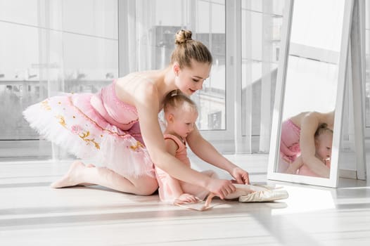 Two ballerinas, mother and daughter sitting on the twine in front of large windows in studio. Little ballerina girl and her teacher doing the split. Little girl stretching with her mother