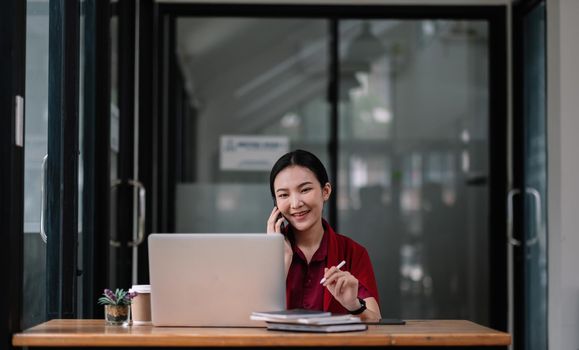 Portrait young asian businesswoman beautiful charming smiling and talking on the mobile phone in the office. female working with laptop computer.