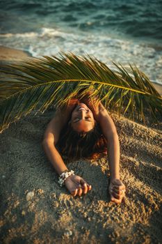 Shot of an attractive young woman is lying on the tropical sandy beach covered with palm tree leaf.