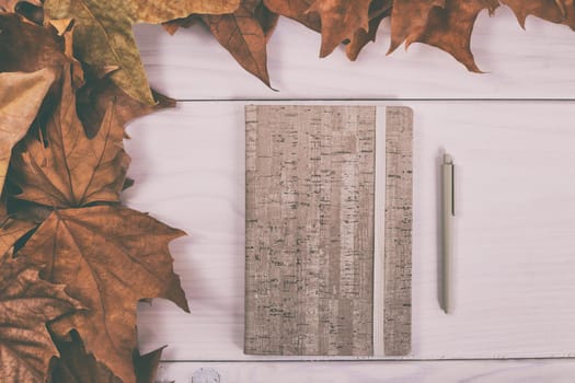 Notebook and pen on wooden table with autumn leaves.Image is intentionally toned.