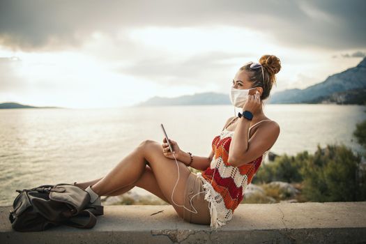 Shot of a happy young woman with protective mask spending time on seaside and listening music from her smartphone during exploring a Mediterranean at corona pandemic. 